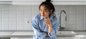 A Woman Stands In Her Kitchen With A Glass Of Water Contemplating Medical Detox Options.