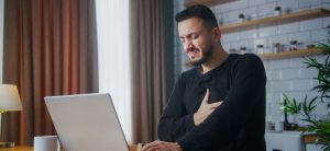 Man Using Laptop Grimaces As He Grabs His Chest Area.